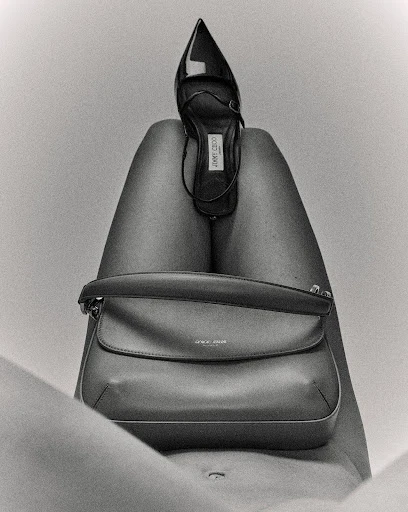 A black and white shot of a woman's legs with a black designer high heel placed on the upper thighs and a handbag on the lower thighs.