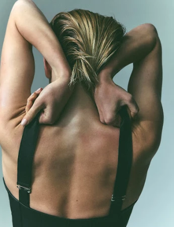 Rear view of a person with blonde hair holding their hands behind their neck, wearing a black garment against a grey background.