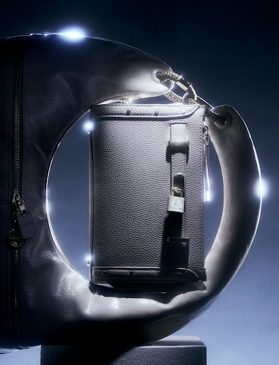 Fashion shot showcases an elegant gray leather handbag with a sculptural handle and metallic accents, set against a dark studio backdrop.