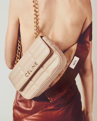 Tan Celine bag with a gold chain on a model in a leather skirt. Luxury fashion accessory advertisement with a clean background.