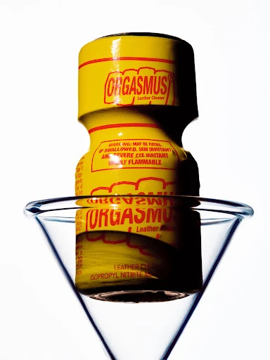 Yellow bottle of 'Orgasmus' leather cleaner inside a clear martini glass against a bright white background.
