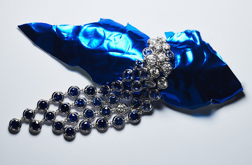 Close-up of a jeweled silver necklace with blue and clear gems, artfully displayed on a piece of crumpled blue foil, studio lighting.