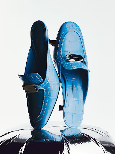 Close-up product shot of bright blue embossed leather mules resting vertically on a shiny silver reflective surface