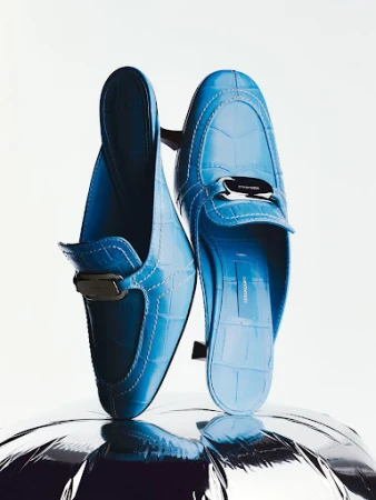 Close-up product shot of bright blue embossed leather mules resting vertically on a shiny silver reflective surface