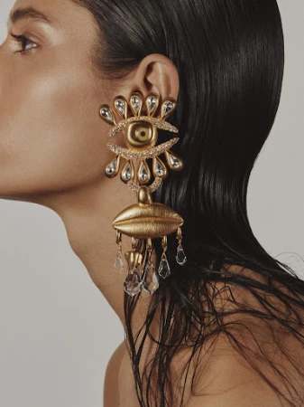 Close-up profile of a woman wearing an oversized ornate gold statement earring featuring an eye and lips design.