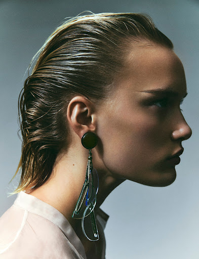 Profile portrait of a woman with wet blonde hair and a large modern earring against a neutral studio backdrop in a beauty editorial.