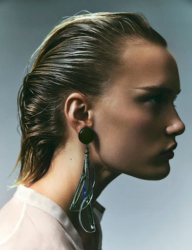 Profile portrait of a woman with wet blonde hair and a large modern earring against a neutral studio backdrop in a beauty editorial.
