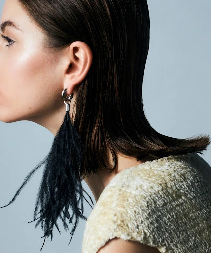 Profile view of a fair-skinned young woman with dark hair wearing a beige top and large, feathery black earring against a gray background.