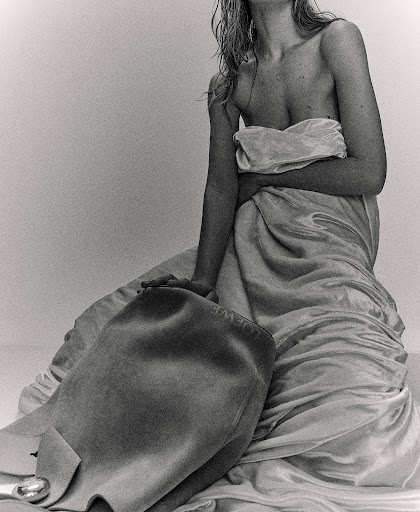 Black and white studio shot of a partially nude adult female with wet blonde hair draped in fabric, holding a leather handbag.