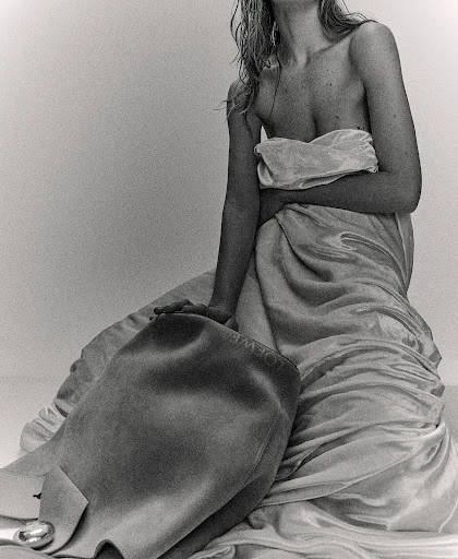 Black and white studio shot of a partially nude adult female with wet blonde hair draped in fabric, holding a leather handbag.