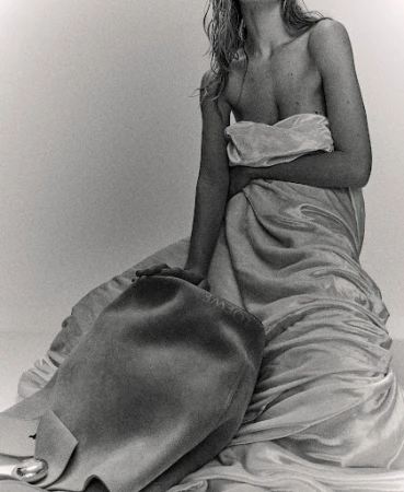 Black and white studio shot of a partially nude adult female with wet blonde hair draped in fabric, holding a leather handbag.