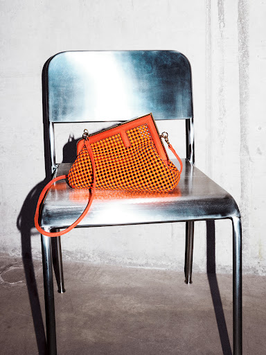 Orange and black woven handbag with bright orange trim and strap, laying on a silver metal chair against a white wall.