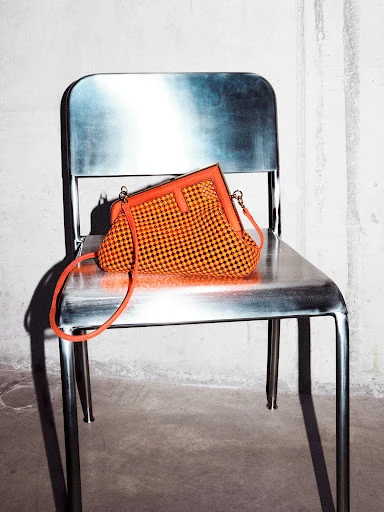Orange and black woven handbag with bright orange trim and strap, laying on a silver metal chair against a white wall.