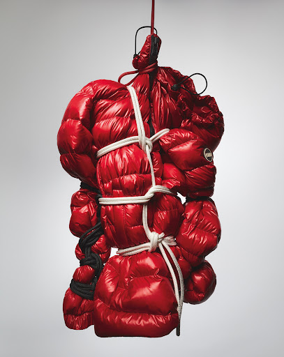Abstract studio shot: glossy red puffer jacket pieces bundled with white and black rope, suspended on metal hanger against pale grey.