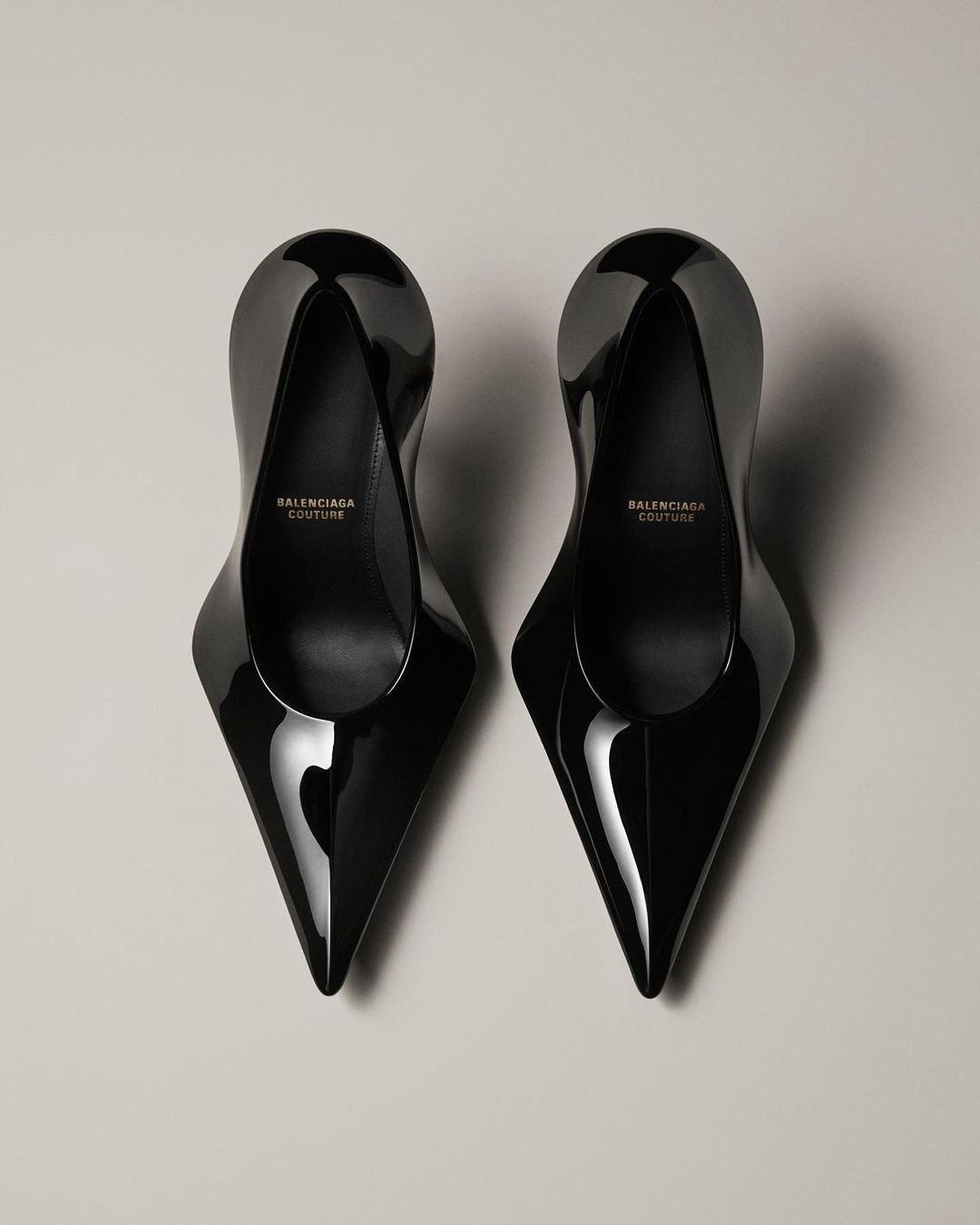 Pair of glossy black Balenciaga Couture high-heel shoes with pointed toes, displayed on a grey surface. Fashion product shot.