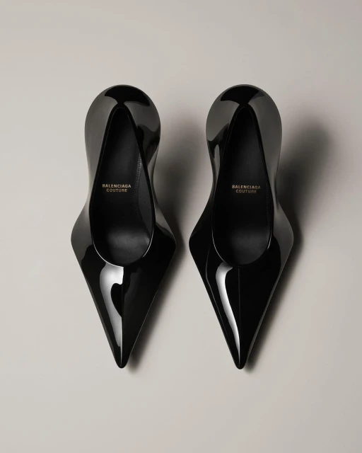 Pair of glossy black Balenciaga Couture high-heel shoes with pointed toes, displayed on a grey surface. Fashion product shot.