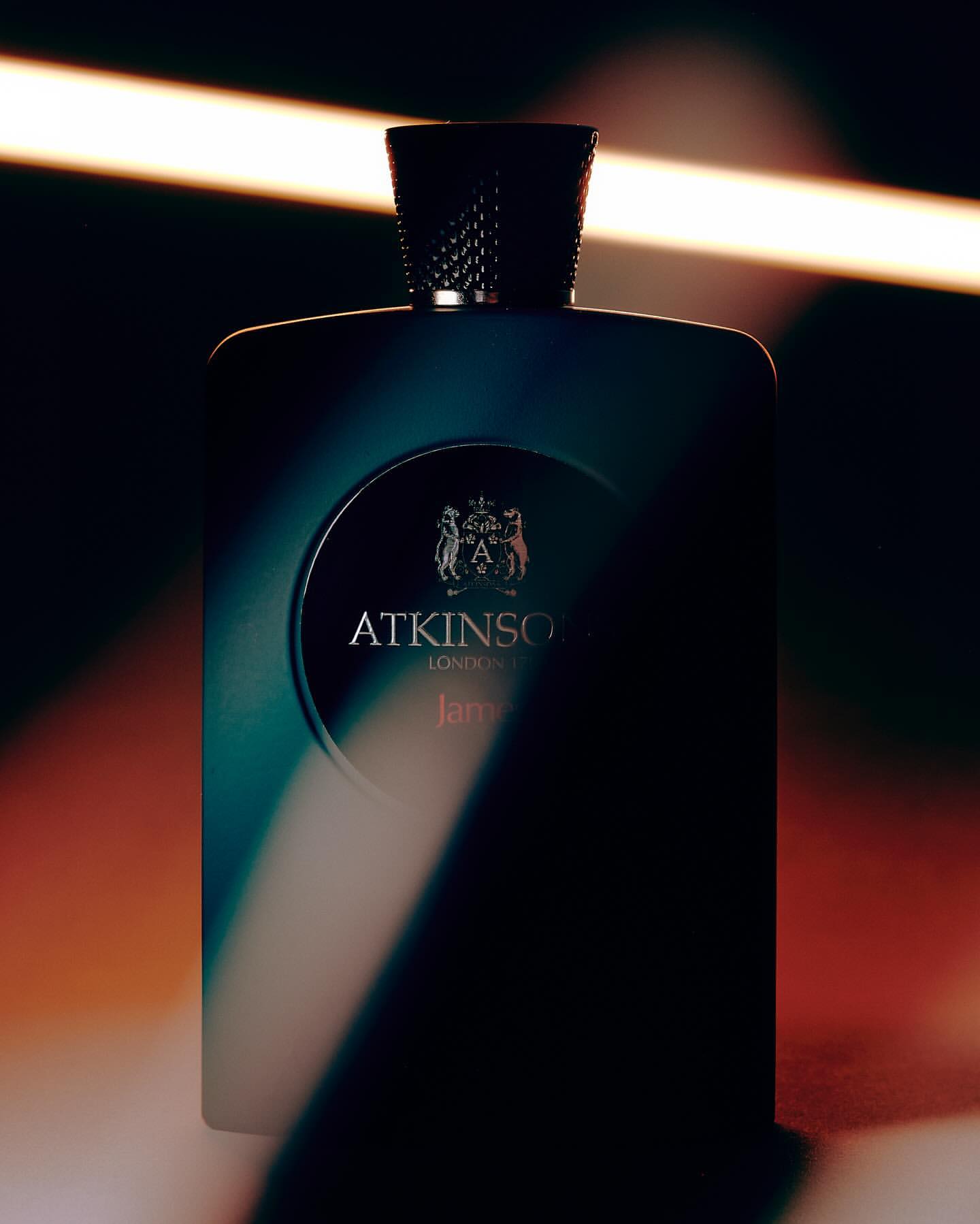 Close-up shot of an elegant, dark teal Atkinson's fragrance bottle against a dimly lit backdrop with warm light streaks.