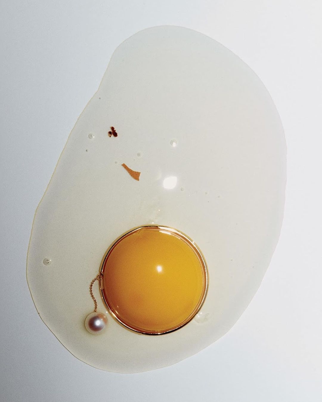Abstract food photo of egg on a white surface featuring jewelry. Visuals include a cracked raw egg, pearl necklace, and gold ring.