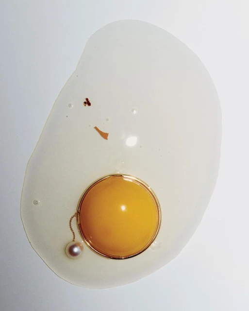 Abstract food photo of egg on a white surface featuring jewelry. Visuals include a cracked raw egg, pearl necklace, and gold ring.