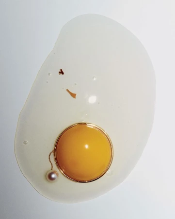 Abstract food photo of egg on a white surface featuring jewelry. Visuals include a cracked raw egg, pearl necklace, and gold ring.
