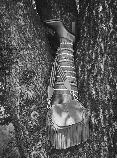 Black and white outdoor shot of a fringed handbag and patterned legwear with a heeled boot, protruding from a tree trunk.