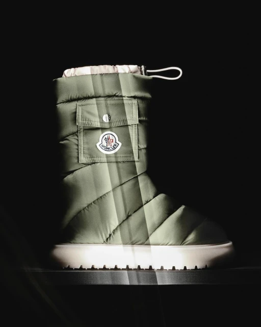 Olive green quilted boot with white sole and Moncler logo detail, captured in a dark, spotlighted setting. Product shot.