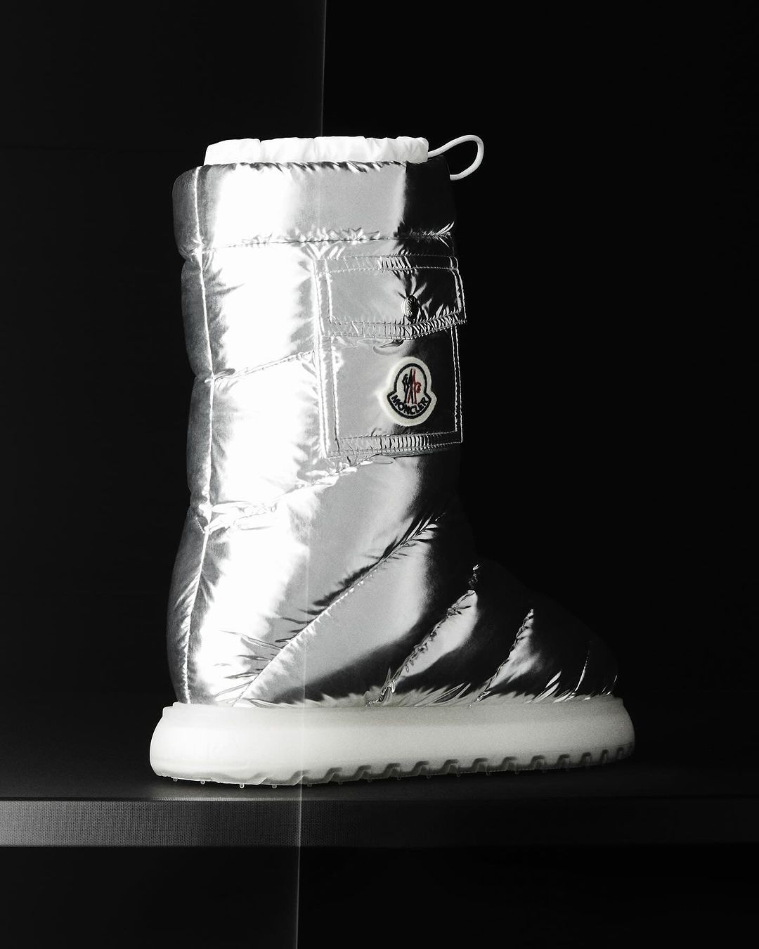 Puffy silver metallic boot by Moncler with white sole, photographed indoors on a black surface against a dark background. Fashion.