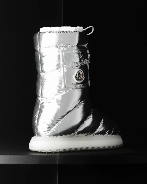 Puffy silver metallic boot by Moncler with white sole, photographed indoors on a black surface against a dark background. Fashion.