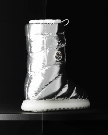 Puffy silver metallic boot by Moncler with white sole, photographed indoors on a black surface against a dark background. Fashion.