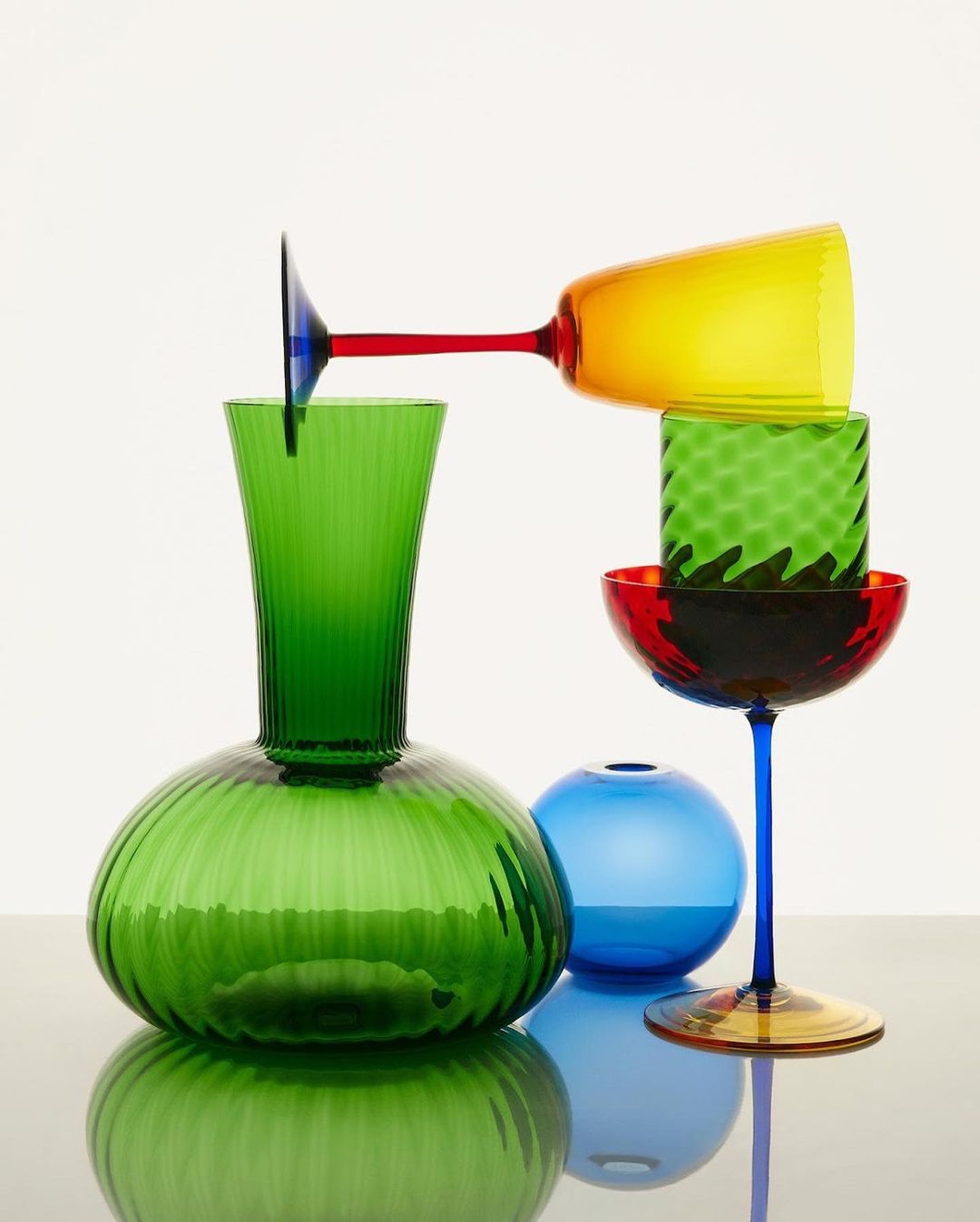 Still life of colorful glassware including vases and stemmed glasses in green, blue, red, and yellow against a white backdrop.