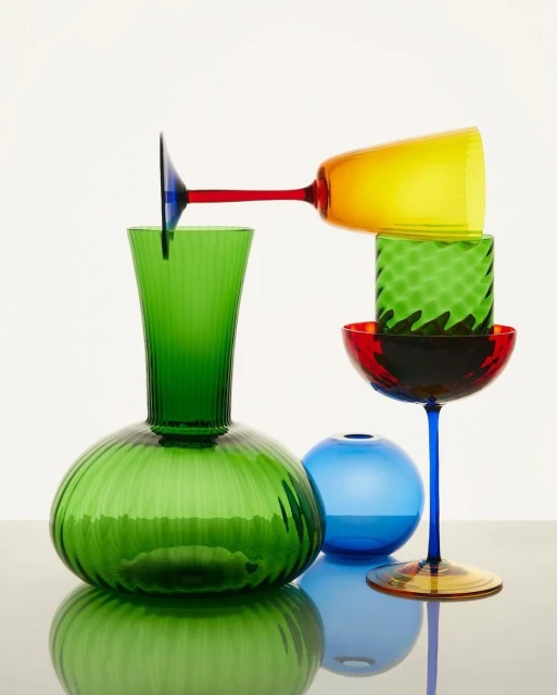 Still life of colorful glassware including vases and stemmed glasses in green, blue, red, and yellow against a white backdrop.