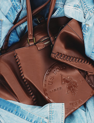Brown US POLO handbag and wallet are partially surrounded by a light blue denim jacket in a studio shot. Fashion product photography.