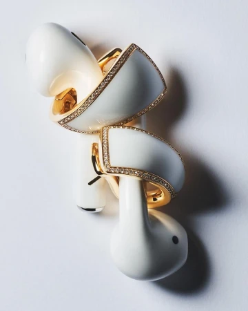 Elevated view of white wireless earbuds featuring a white and gold jewellery wrap around the earpieces, studio lit on a white surface.