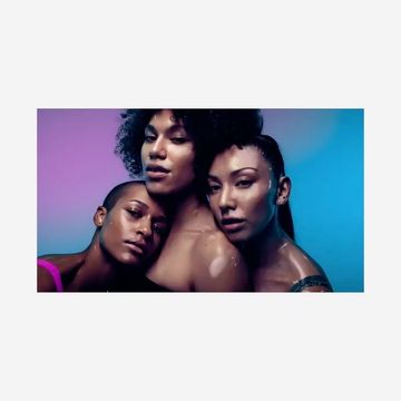 Three women with various skin tones pose together in a studio illuminated by purple and blue lighting, showcasing their bare shoulders and wet skin.