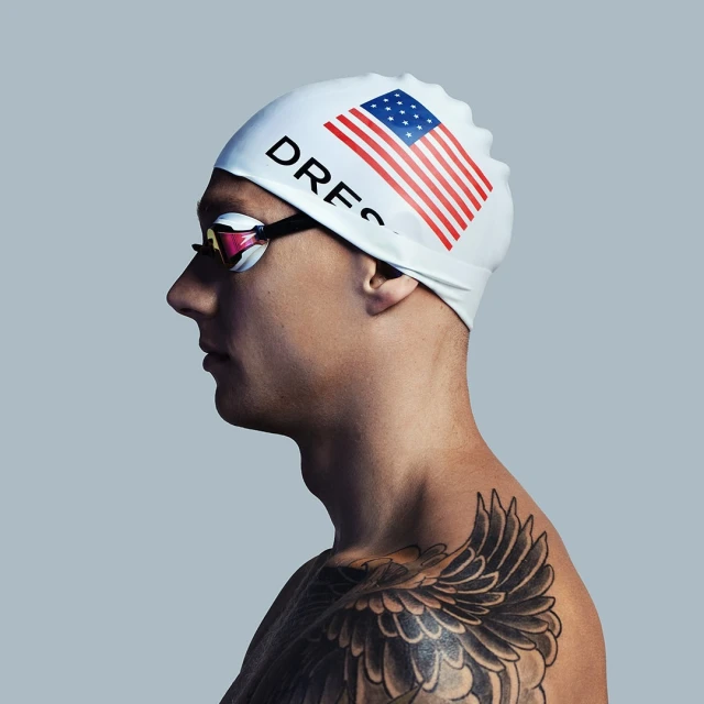 Profile shot of a man in a swim cap with an American flag design and swim goggles, showing a shoulder tattoo against a light gray backdrop.