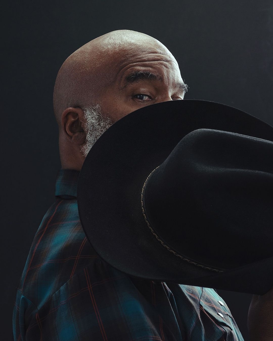 Portrait of an adult African American man wearing a blue plaid shirt and a black cowboy hat against a dark studio backdrop.