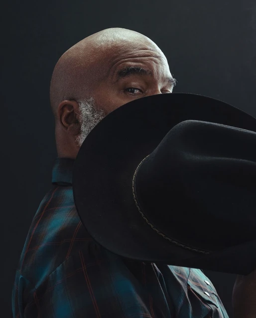 Portrait of an adult African American man wearing a blue plaid shirt and a black cowboy hat against a dark studio backdrop.