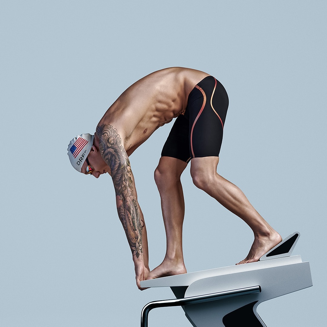 Muscular swimmer wearing a cap and swim trunks on a starting block, leaning forward and preparing to dive, against a light blue backdrop.