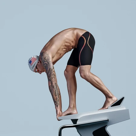 Muscular swimmer wearing a cap and swim trunks on a starting block, leaning forward and preparing to dive, against a light blue backdrop.