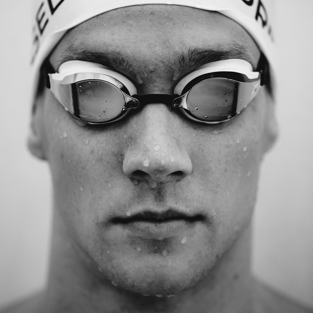 Close-up black and white portrait of a swimmer wearing goggles and a swim cap, face wet with water droplets against a plain background.