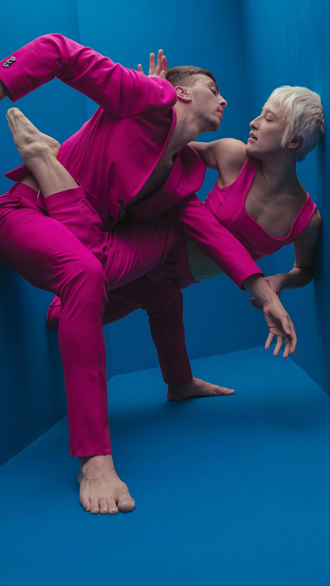 Two people in a dance pose, wearing fuchsia matching outfits, a man in a suit and a woman in a tank top, with a blue background.