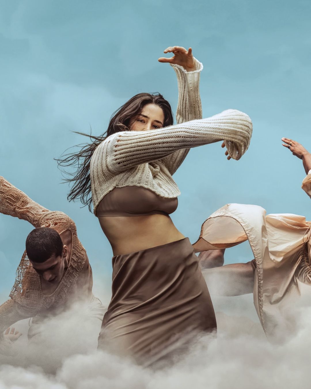 Fashionable woman dances in ethereal scene with two other people in cream tones against a soft sky backdrop with cloud-like effects.