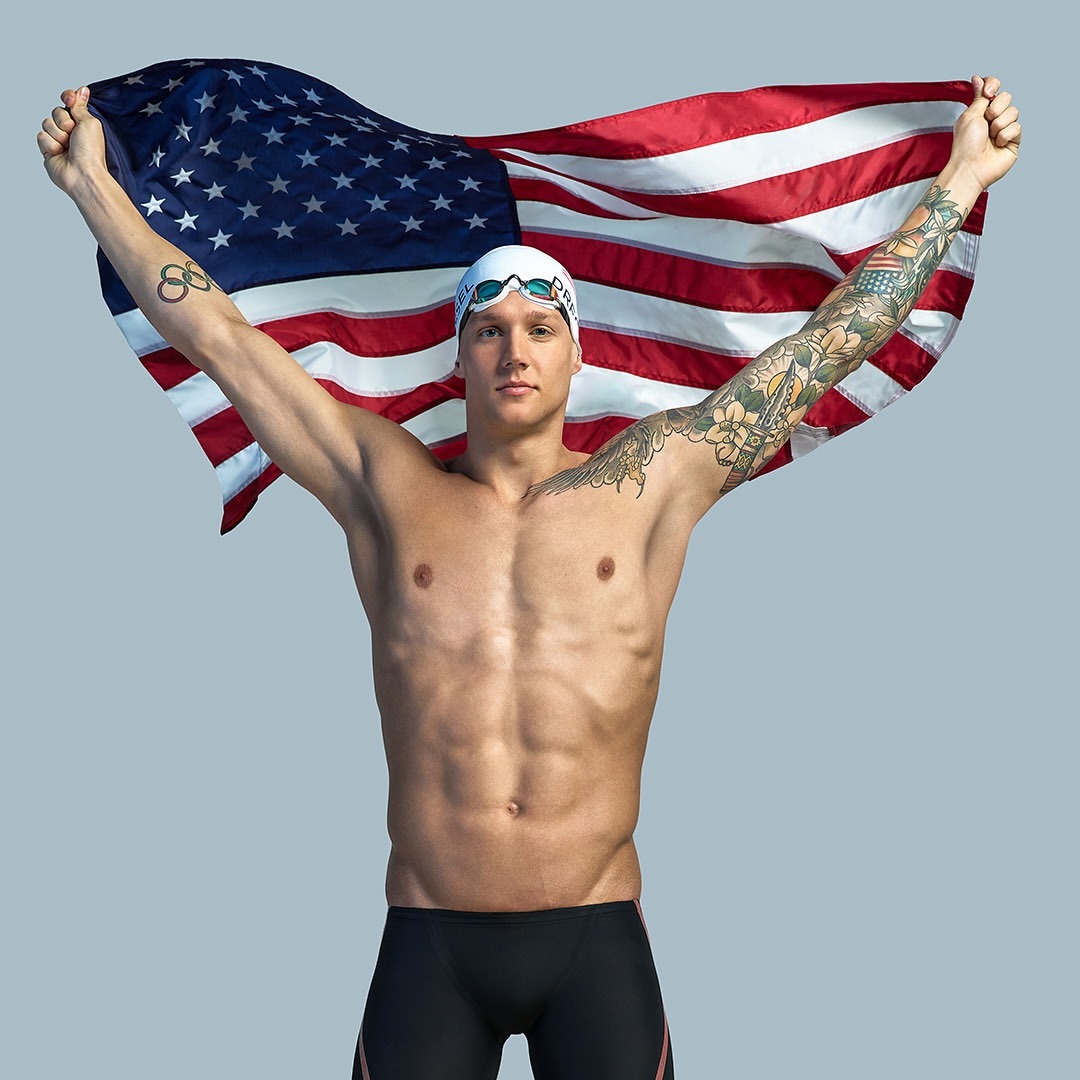 Muscular swimmer posing shirtless with an American flag held above his head in a studio environment, wearing swim cap and goggles.