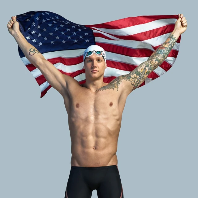 Muscular swimmer posing shirtless with an American flag held above his head in a studio environment, wearing swim cap and goggles.