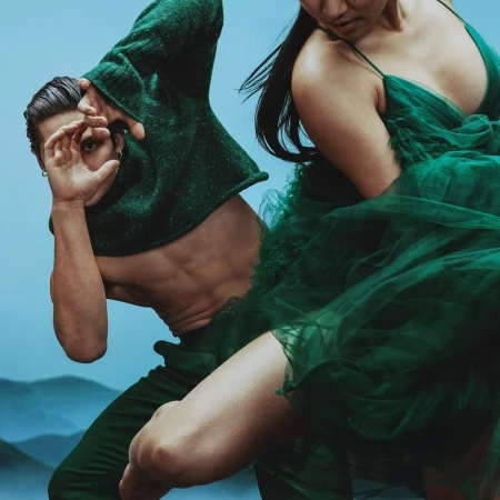 Two people in motion, an adult male making a frame with his hands and an adult female wearing a green dress, against a blue background.