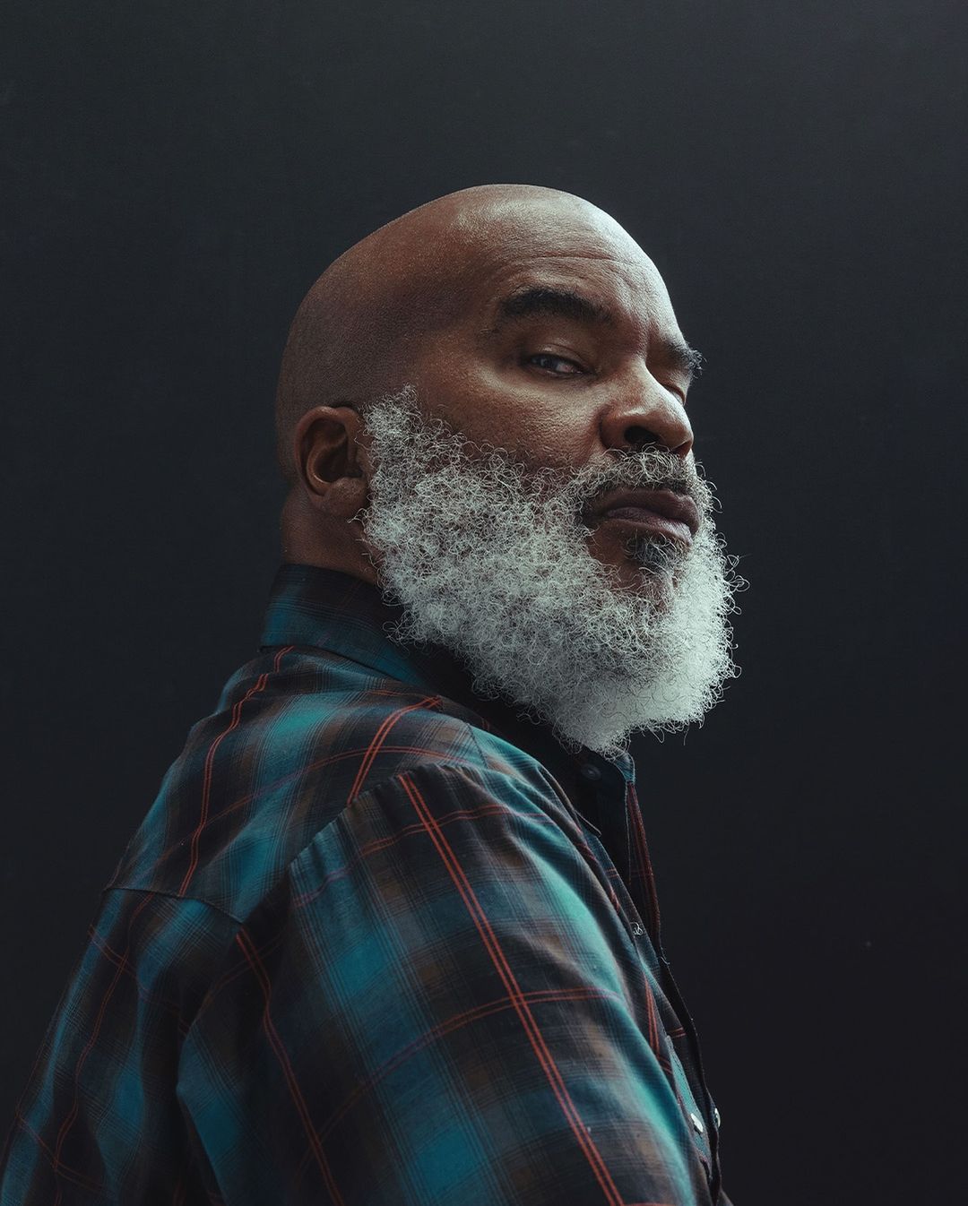 Portrait of an adult Black man with a bald head and white beard looking over his shoulder while wearing a blue and red plaid shirt.