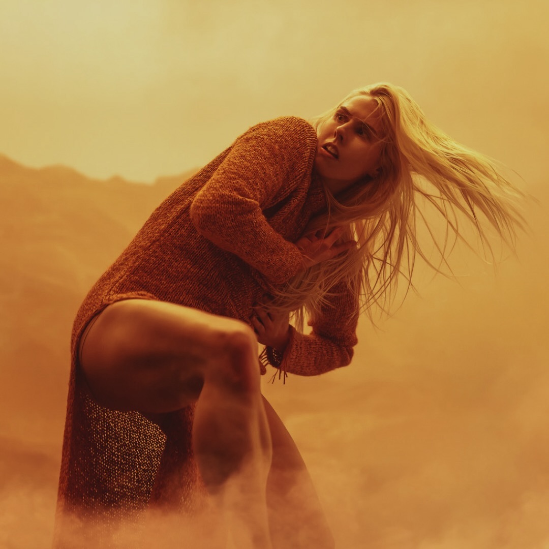 Woman with blonde hair in orange sweater dress in dusty landscape, leaning forward with worried expression, captured in eye-level shot.