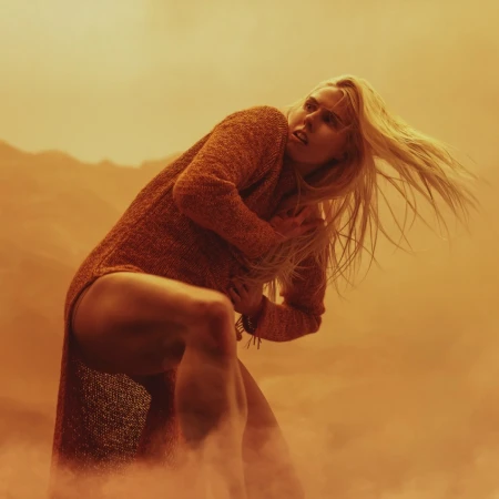 Woman with blonde hair in orange sweater dress in dusty landscape, leaning forward with worried expression, captured in eye-level shot.