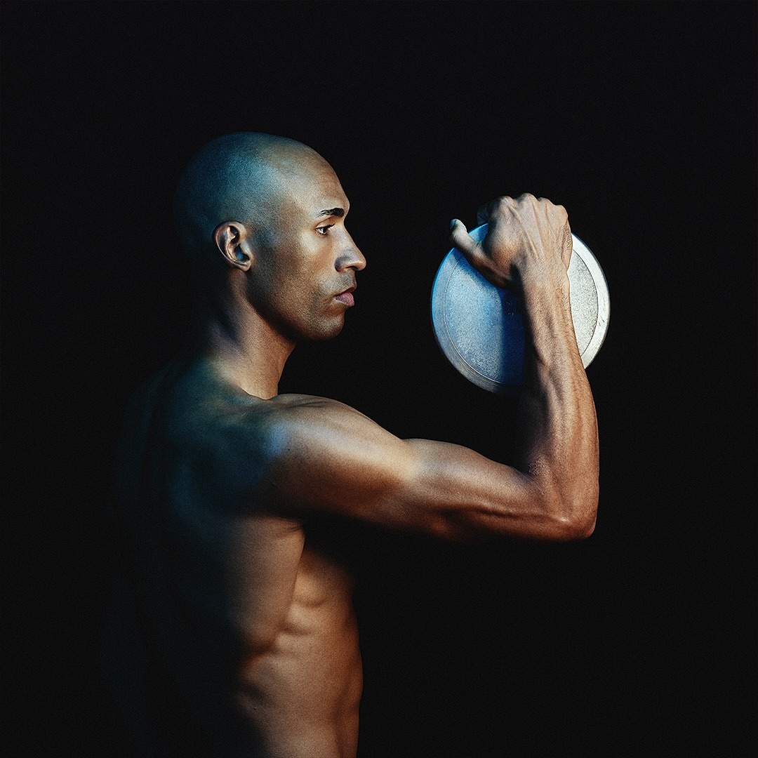 Shirtless man with dark skin holds a grey flying disc in front of a black background in a dramatic studio shot. Sport and fitness concept.