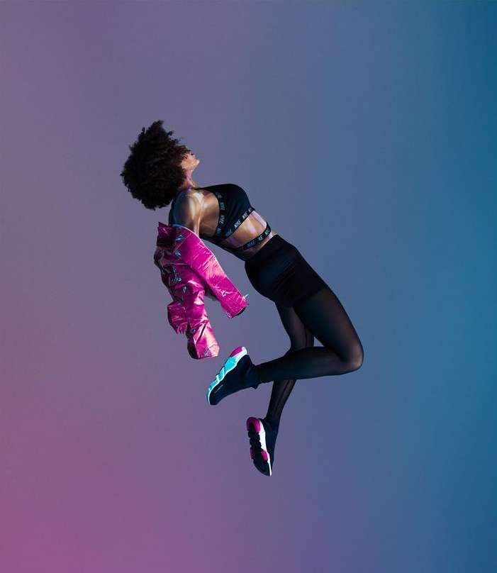 Athletic woman in sports bra and shorts, mid-air with pink jacket, against colorful gradient backdrop, showcasing fitness and style.